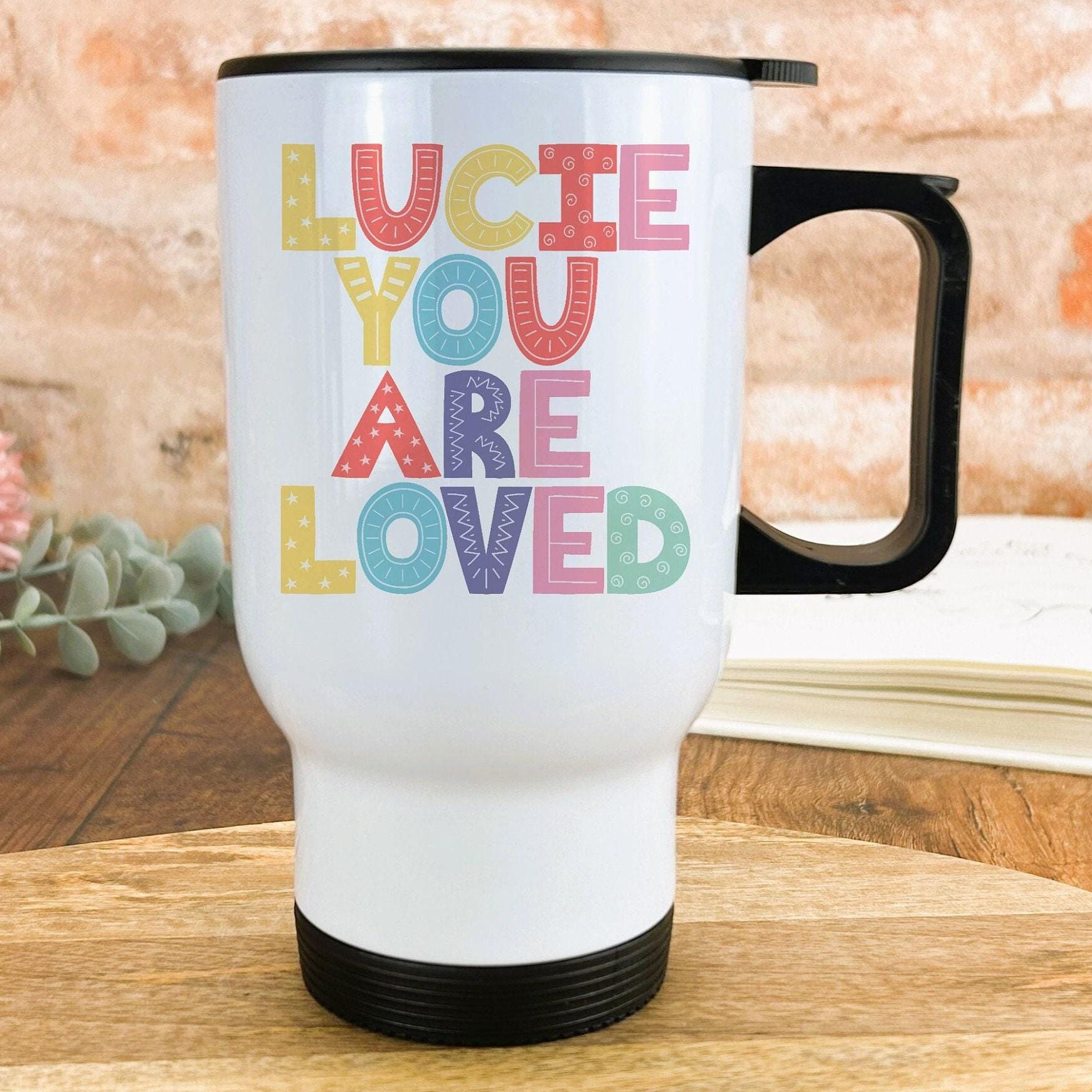 A white travel mug with colorful text that says "LUCIE YOU ARE LOVED" on it.