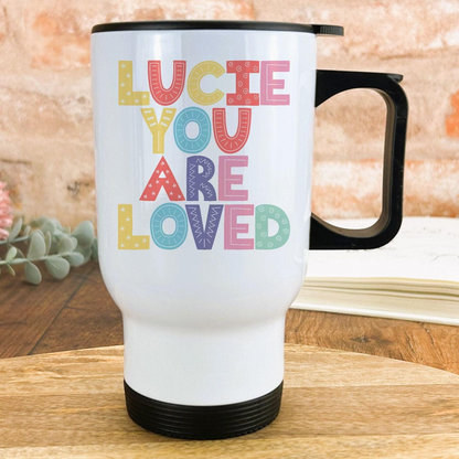 A white travel mug with colorful text that says "LUCIE YOU ARE LOVED" on it.