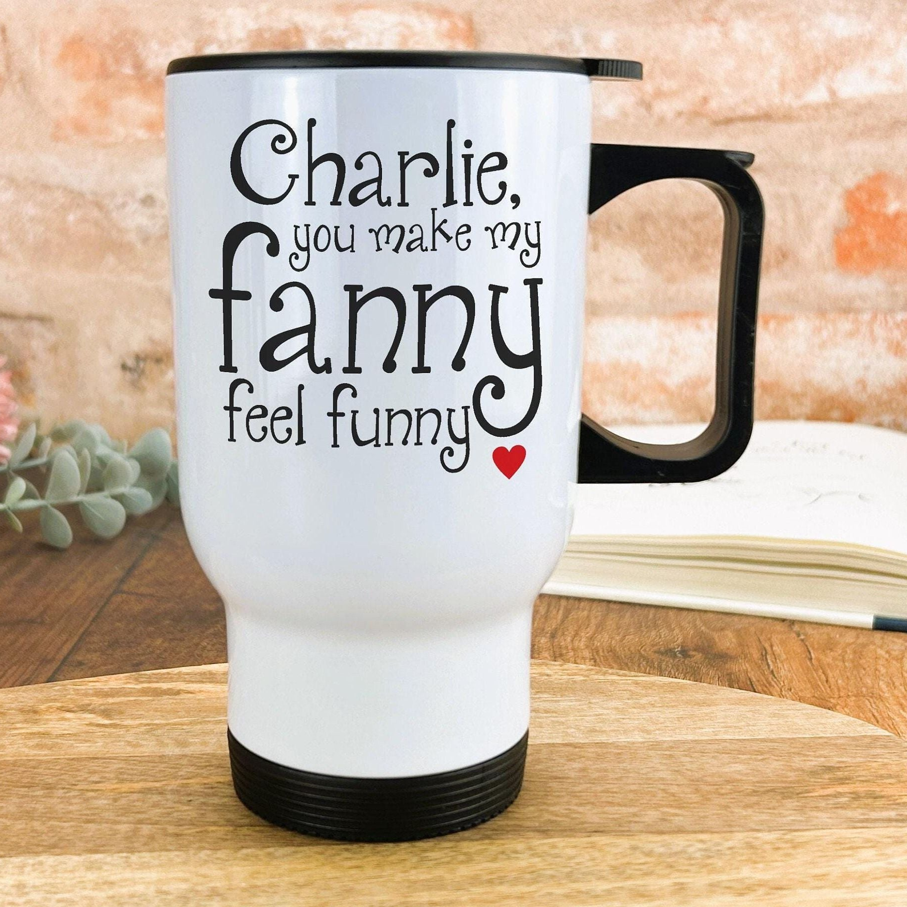 A white travel mug with black text and a red heart on the side, placed on a wooden surface against a brick wall background.