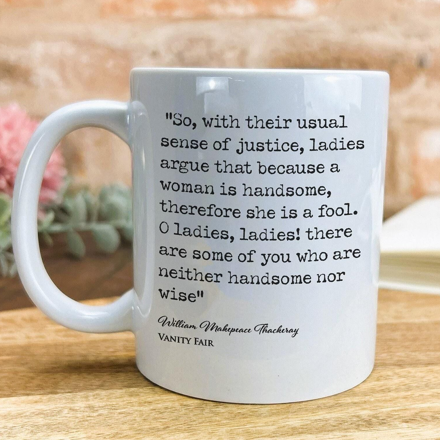A white ceramic mug with a quote from William Shakespeare printed on it, placed on a wooden surface.