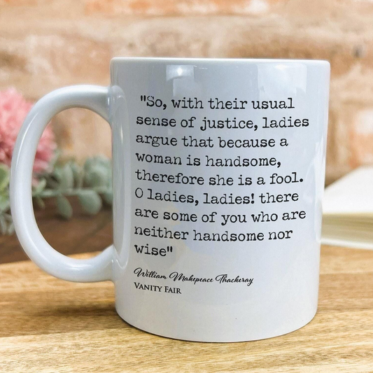 A white ceramic mug with a quote from William Shakespeare printed on it, placed on a wooden surface.