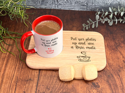 A red and white coffee mug with a message about putting plates up and a Rosie mate, accompanied by a wooden tray with cookies and a plant on a wooden surface.
