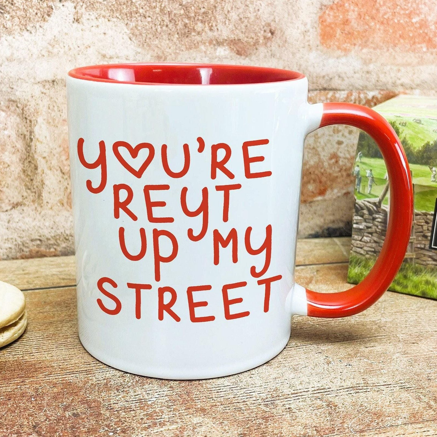 A white mug with a red handle, featuring the text "YOU'RE REYT UP MY STREET" printed on it.