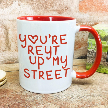 A white mug with a red handle, featuring the text "YOU'RE REYT UP MY STREET" printed on it.