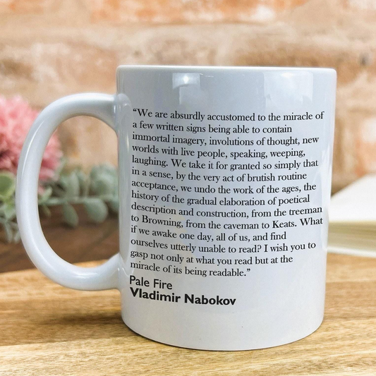 A white ceramic mug with a quote from Vladimir Nabokov printed on it, placed on a wooden surface.