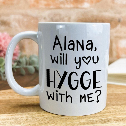 A white ceramic mug with black text that reads "Alana, will you hygge with me?"