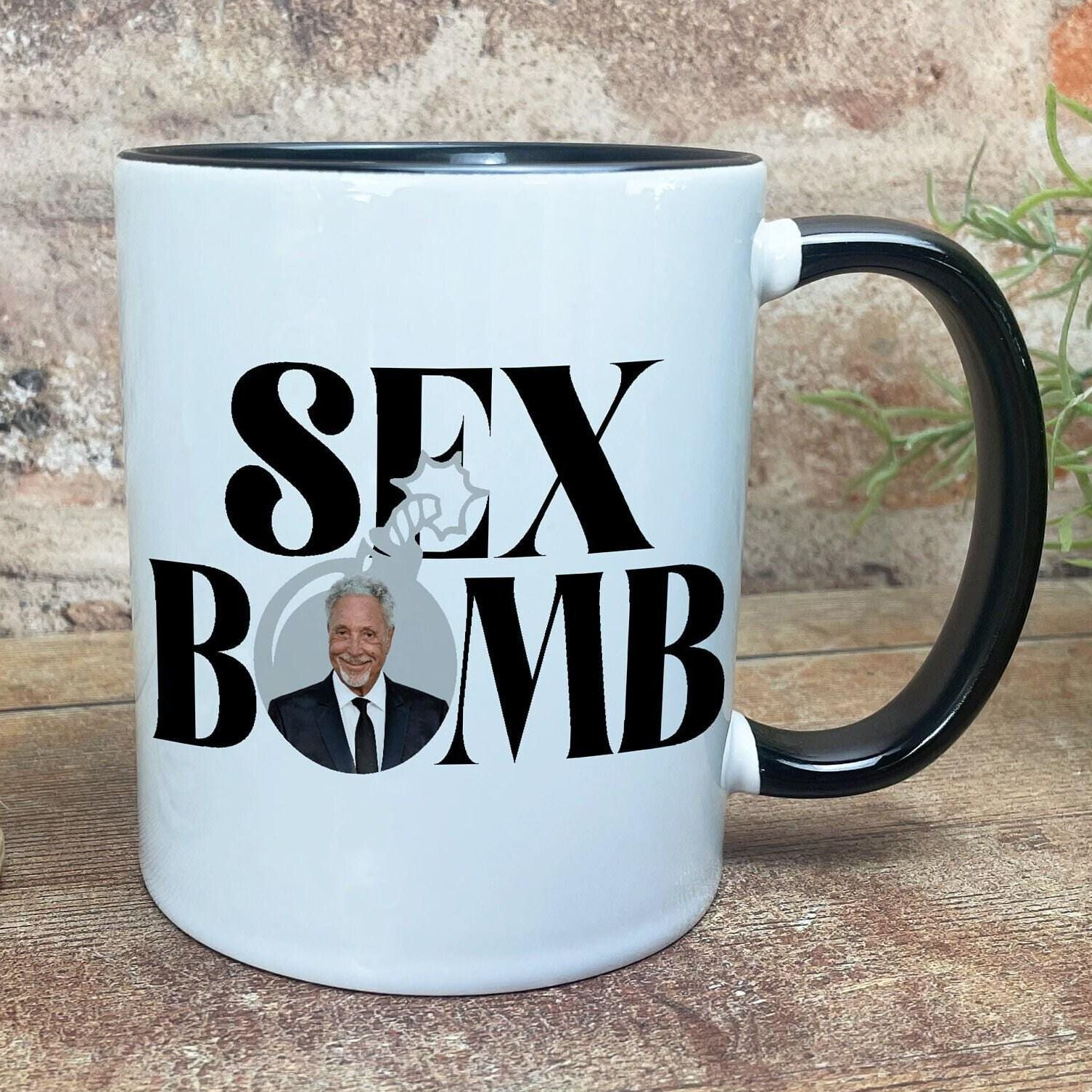 A white ceramic mug with the text "SEX BOMB" printed on it, along with a portrait of a man wearing a suit.