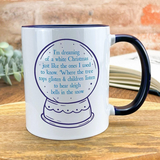 A white mug with a blue handle, featuring a snow globe design and text that reads "I'm dreaming of a white Christmas just like the ones I used to know. Where the tree tops glisten & children listen to hear sleigh bells in the snow".
