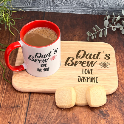 A red and white coffee mug with the text "Dad's Brew" and "Love Jasmine" printed on it, placed on a wooden cutting board. There are also two cookies and some greenery in the background.