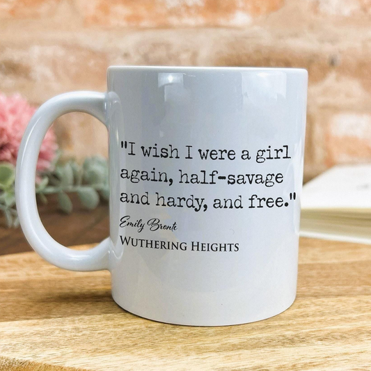 A white ceramic mug with a quote from Emily Brontë printed on it, placed on a wooden surface against a brick wall background.