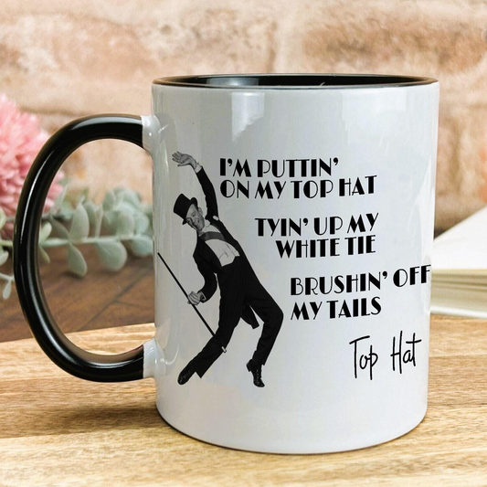 A white mug with a black handle and a black and white illustration of a man dancing. The mug has text that reads "I'm puttingtin' on my top hat, twin' up my white tie, brushin' off my tails, Top Hat".