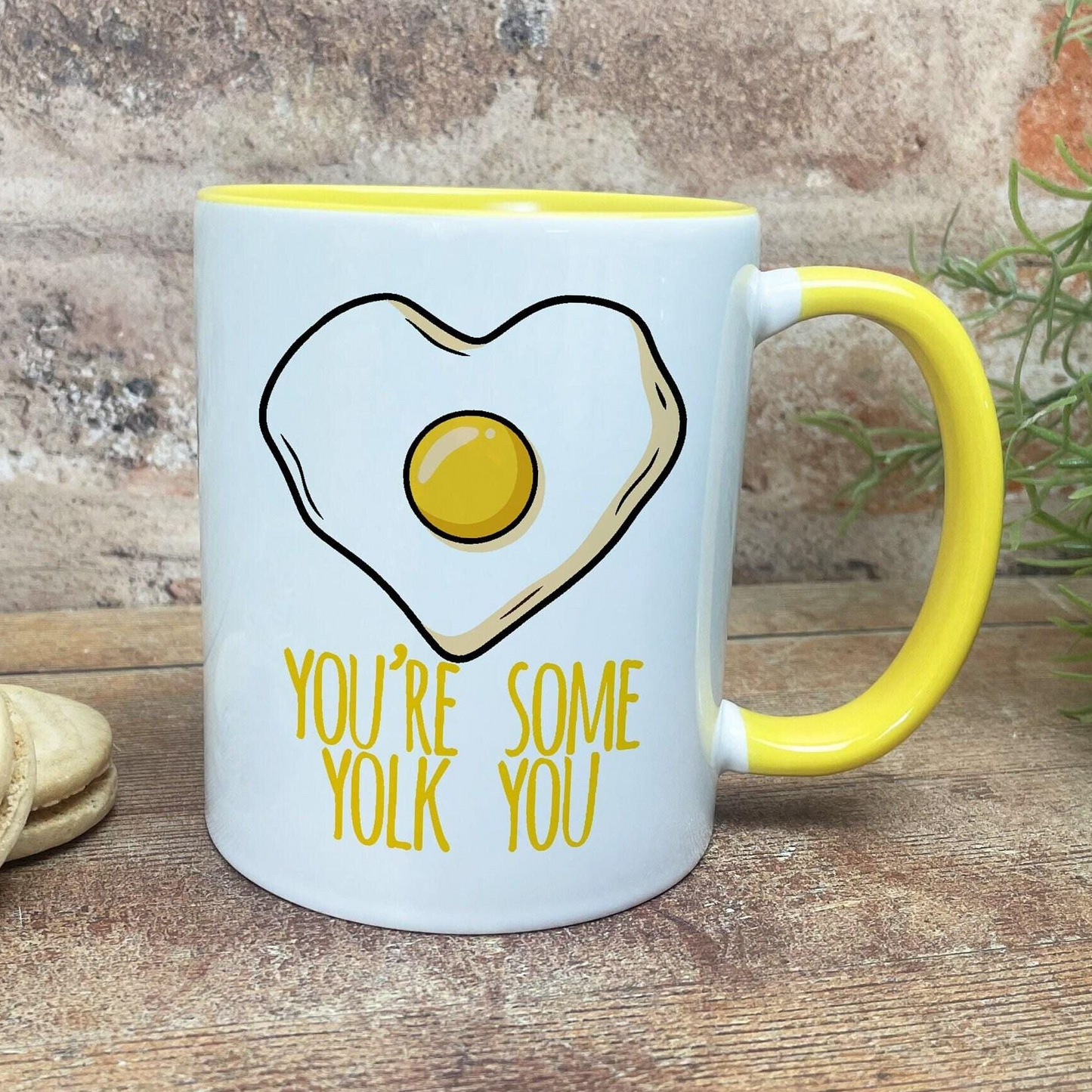 A white mug with a yellow handle, featuring a heart-shaped illustration of an egg and the text "YOU'RE SOME YOLK YOU" printed on it.