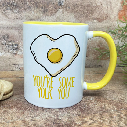 A white mug with a yellow handle, featuring a heart-shaped illustration of an egg and the text "YOU'RE SOME YOLK YOU" printed on it.