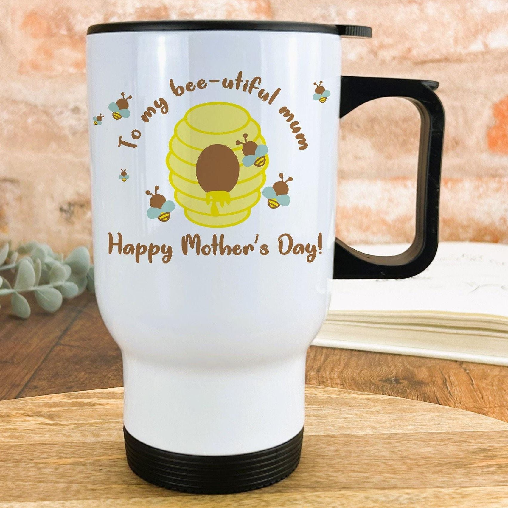 A white travel mug with a black handle, featuring a cartoon image of a honeycomb and the text "To my bee-utiful mum Happy Mother's Day!"