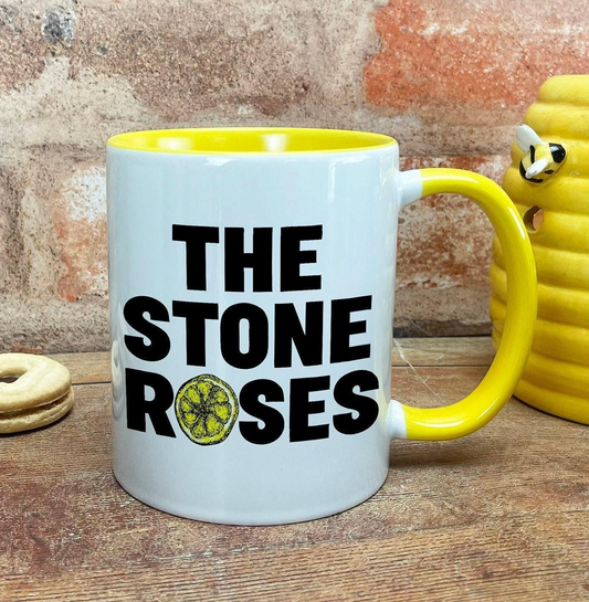 A white and yellow mug with the text "THE STONE ROSSES" printed on it, placed on a wooden surface next to a yellow object.