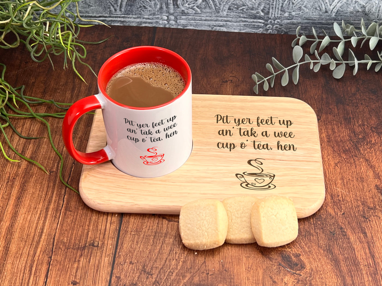 A red and white coffee mug with a humorous message, placed on a wooden tray alongside some cookies, with a plant and greenery in the background.