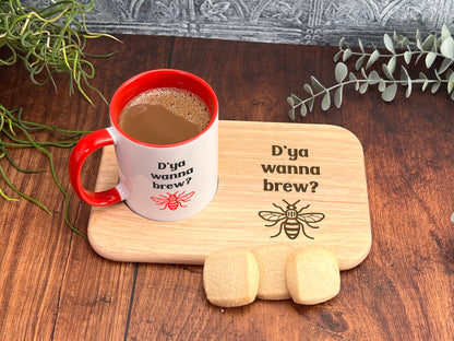 A red and white coffee mug with the text "Dya Wanna brew?" printed on it, placed on a wooden tray with cookies and greenery.