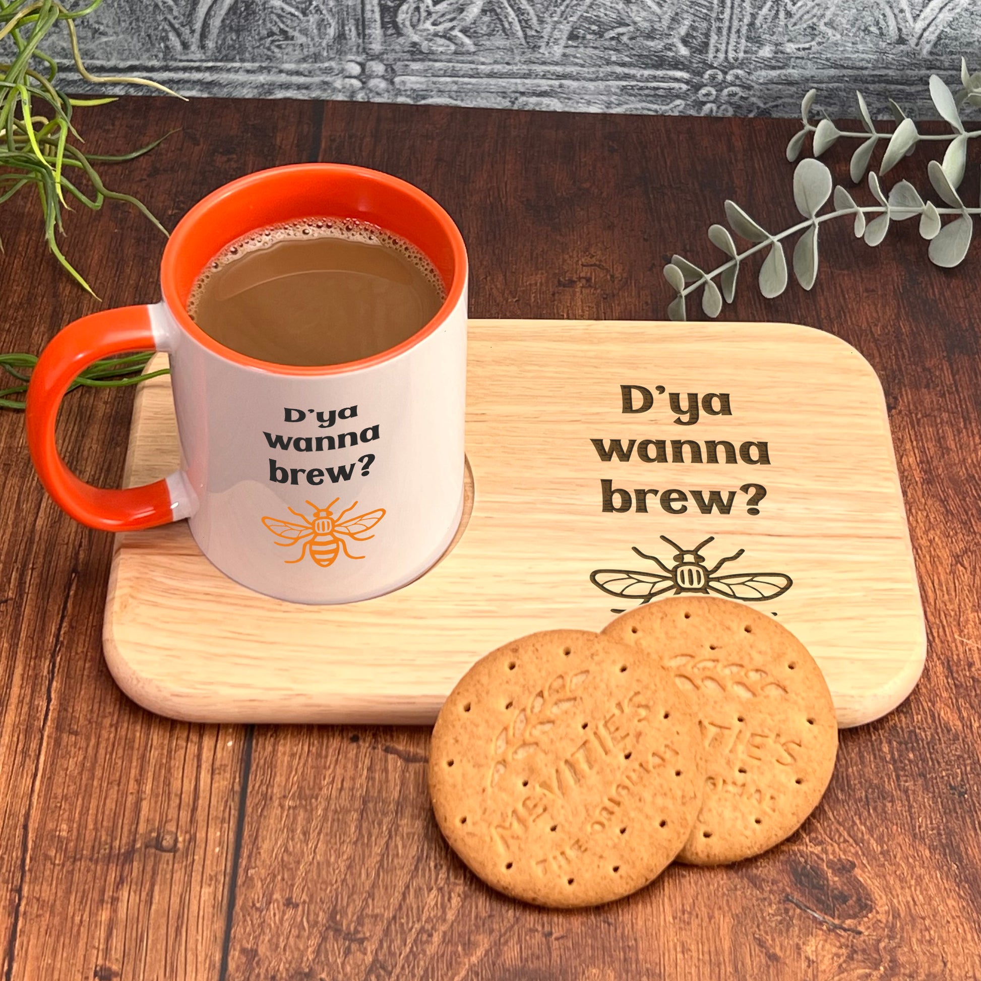 A mug with the text "D'ya wanna brew?" and a wooden tray with cookies and a plant on a wooden surface.