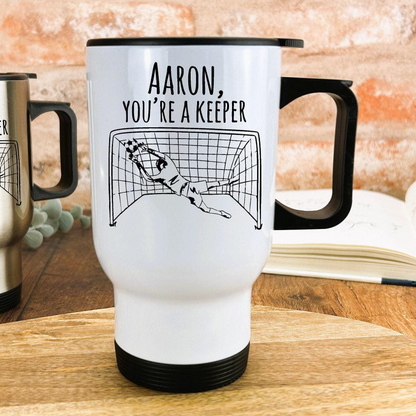 A white travel mug with a black handle and a printed design of a goalkeeper diving to save a soccer ball.