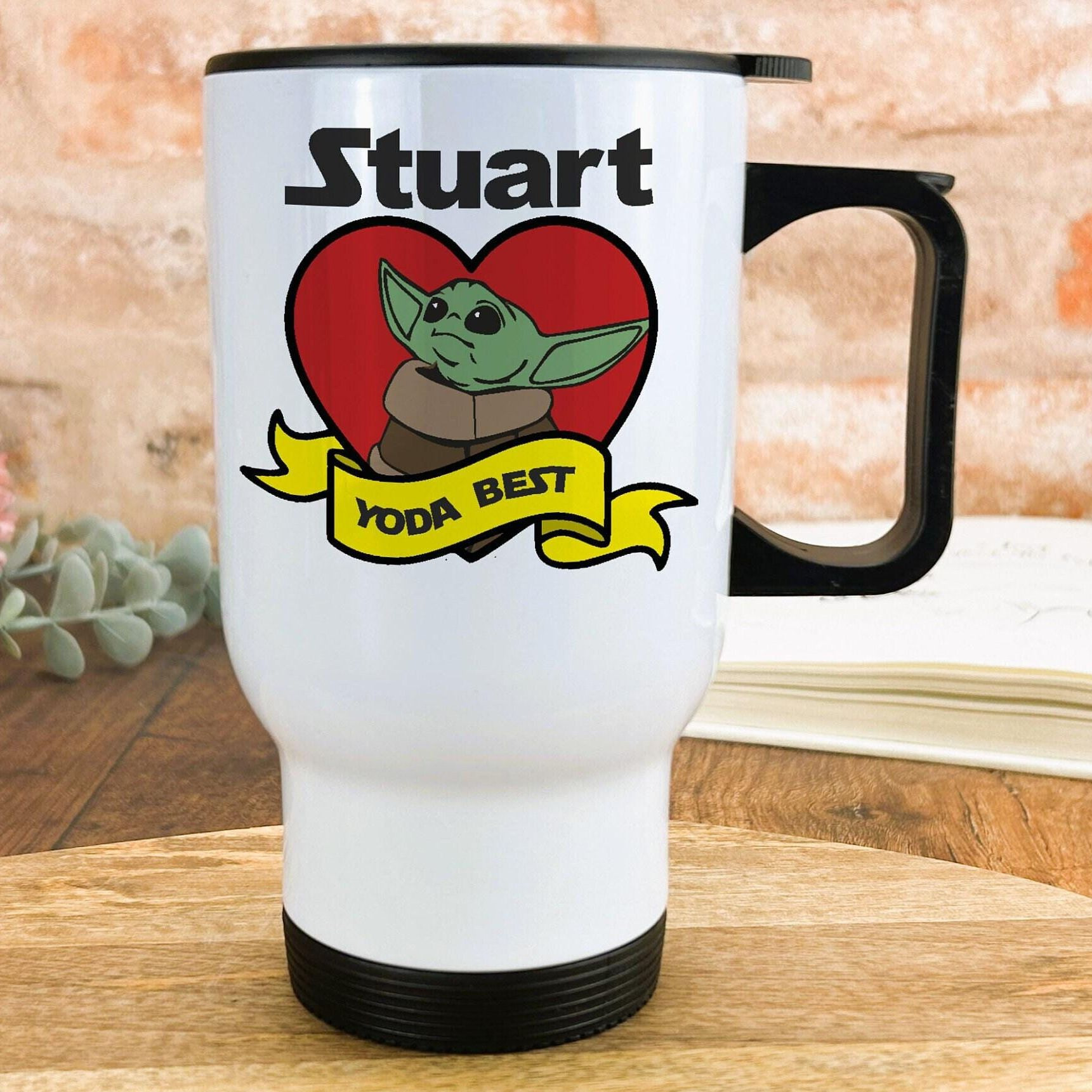 A white travel mug with a black handle, featuring a graphic of a young alien character and the text "YODA BEST" along with a heart symbol.