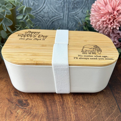A white bento box with a wooden lid, featuring a message engraved on it. The message reads "Happy Mother's Day Love from Step 2. No matter what age, I'll always need you mum." The bento box is placed on a wooden surface, with a pink flower in the background.