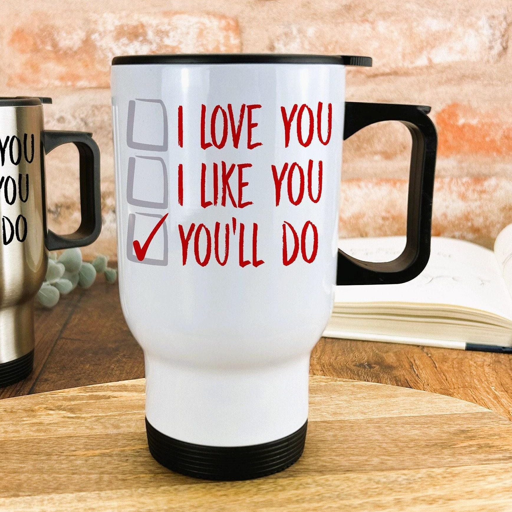 A white travel mug with red text that says "I LOVE YOU I LIKE YOU YOU'LL DO" on it, placed on a wooden surface.