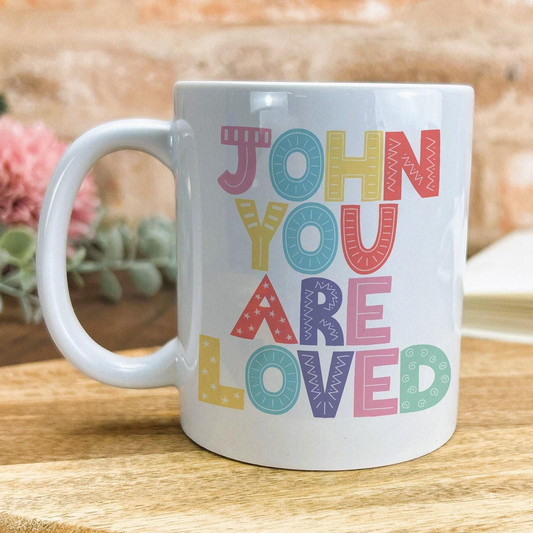 A white ceramic mug with colorful text that says "JOHN YOU ARE LOVED" printed on it.