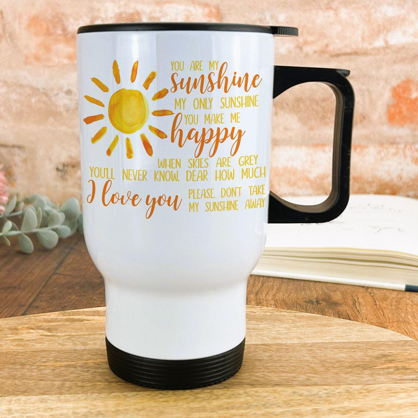 A white travel mug with a black handle, featuring a sun design and text that reads "You are my sunshine, my only sunshine, you make me happy. When skies are grey, I know, dear how much I love you. Please don't take my sunshine away".