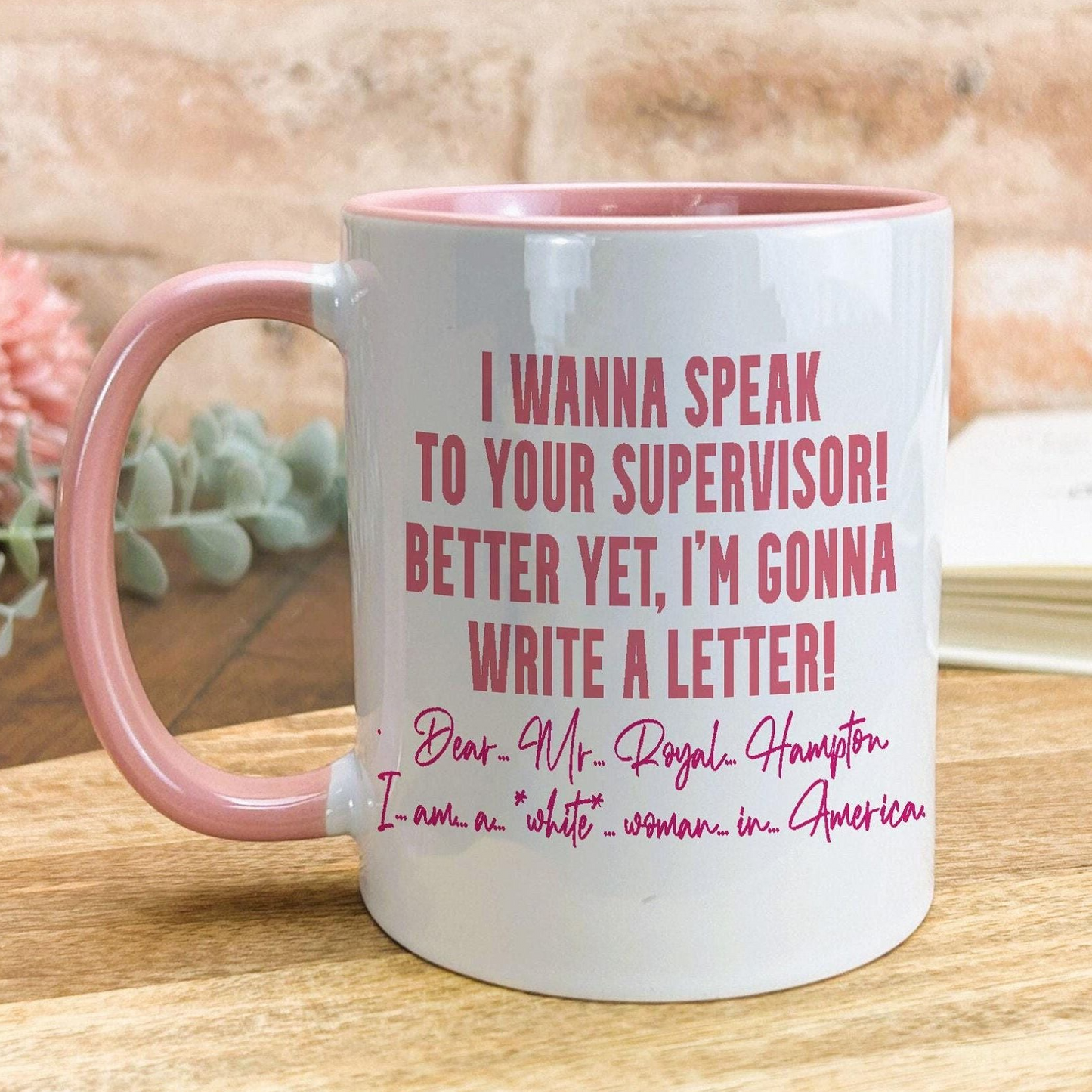 A white mug with a pink handle, featuring a humorous text that reads "I WANNA SPEAK TO YOUR SUPERVISOR! BETTER YET, I'M GONNA WRITE A LETTER!" The mug is placed on a wooden surface, with a pink flower visible in the background.
