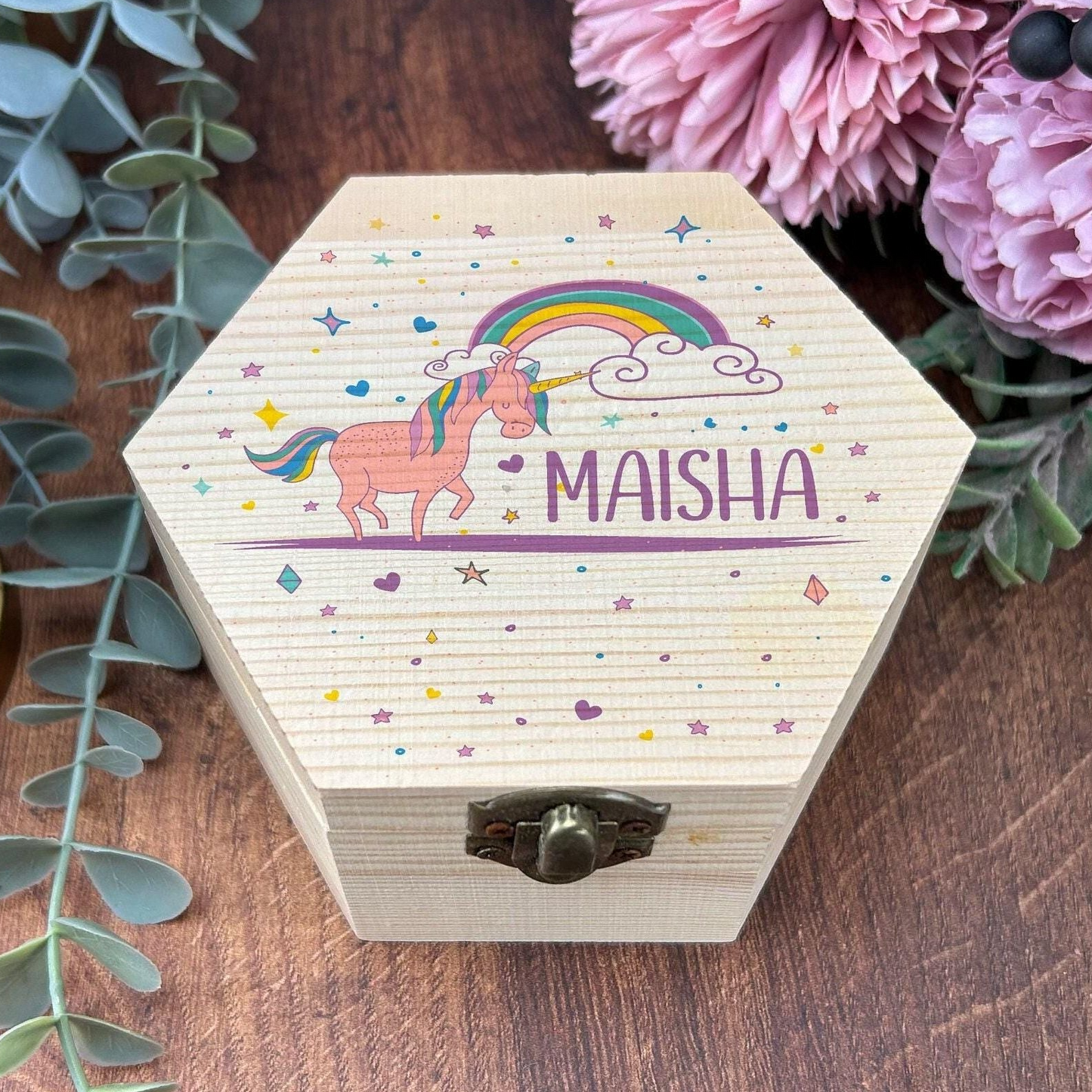 A wooden box with a hexagonal shape, featuring a picture of a unicorn and a rainbow, along with the name "Maisha" written on it. The box is placed on a wooden surface, surrounded by greenery and flowers.