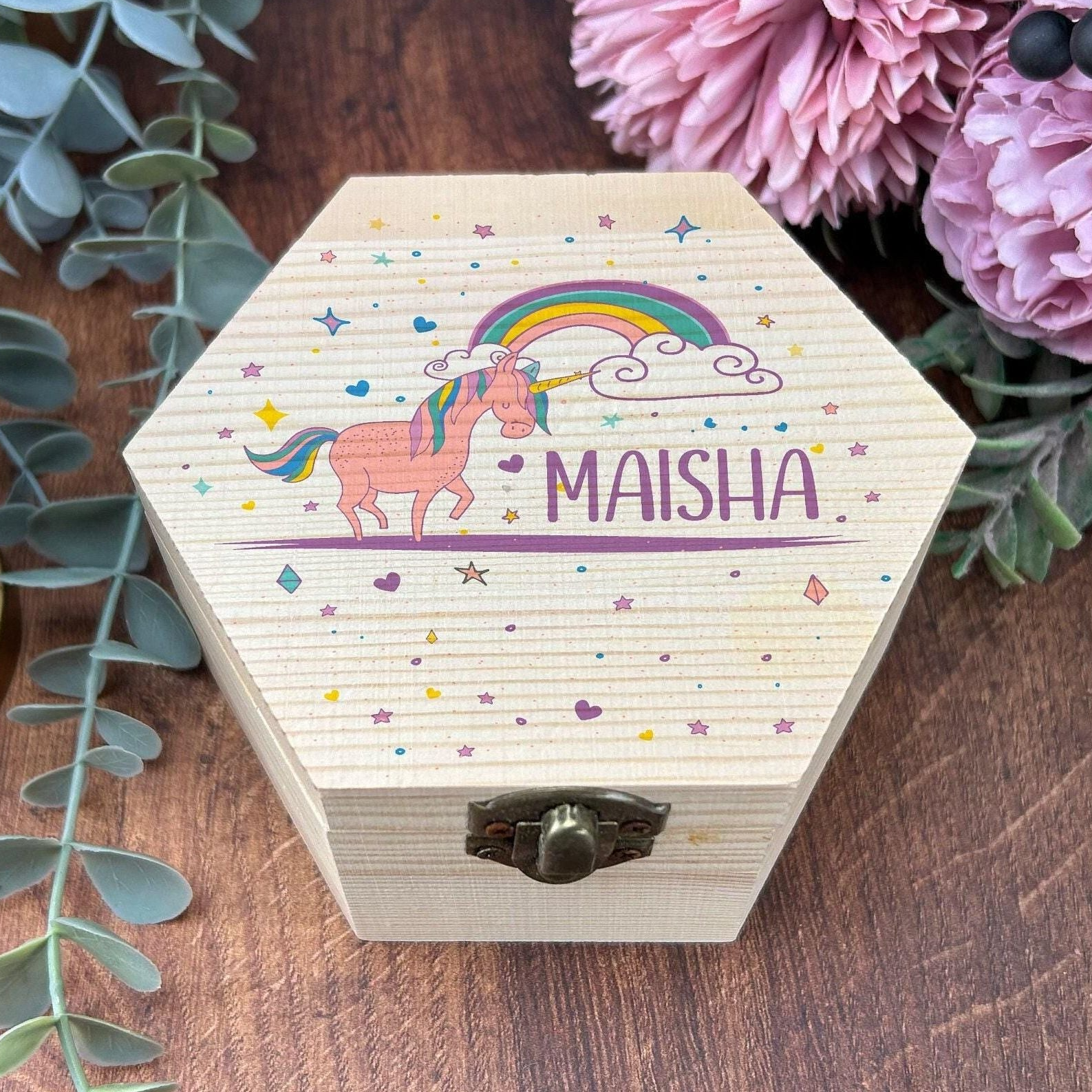 A wooden box with a hexagonal shape, featuring a picture of a unicorn and a rainbow, along with the name "Maisha" written on it. The box is placed on a wooden surface, surrounded by greenery and flowers.