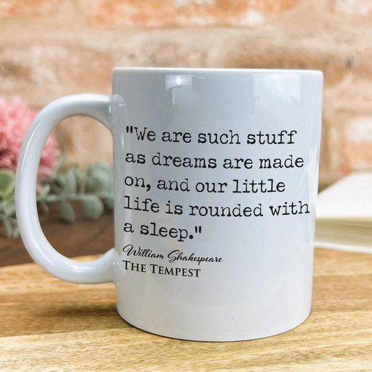 A white ceramic mug with a quote from William Shakespeare printed on it, placed on a wooden surface against a brick wall background.