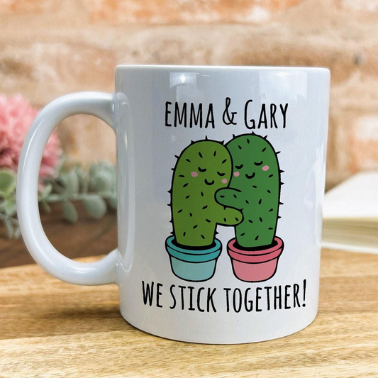 A white ceramic mug with a printed design featuring two cacti hugging each other, accompanied by the text "Emma & Gary We stick together!"