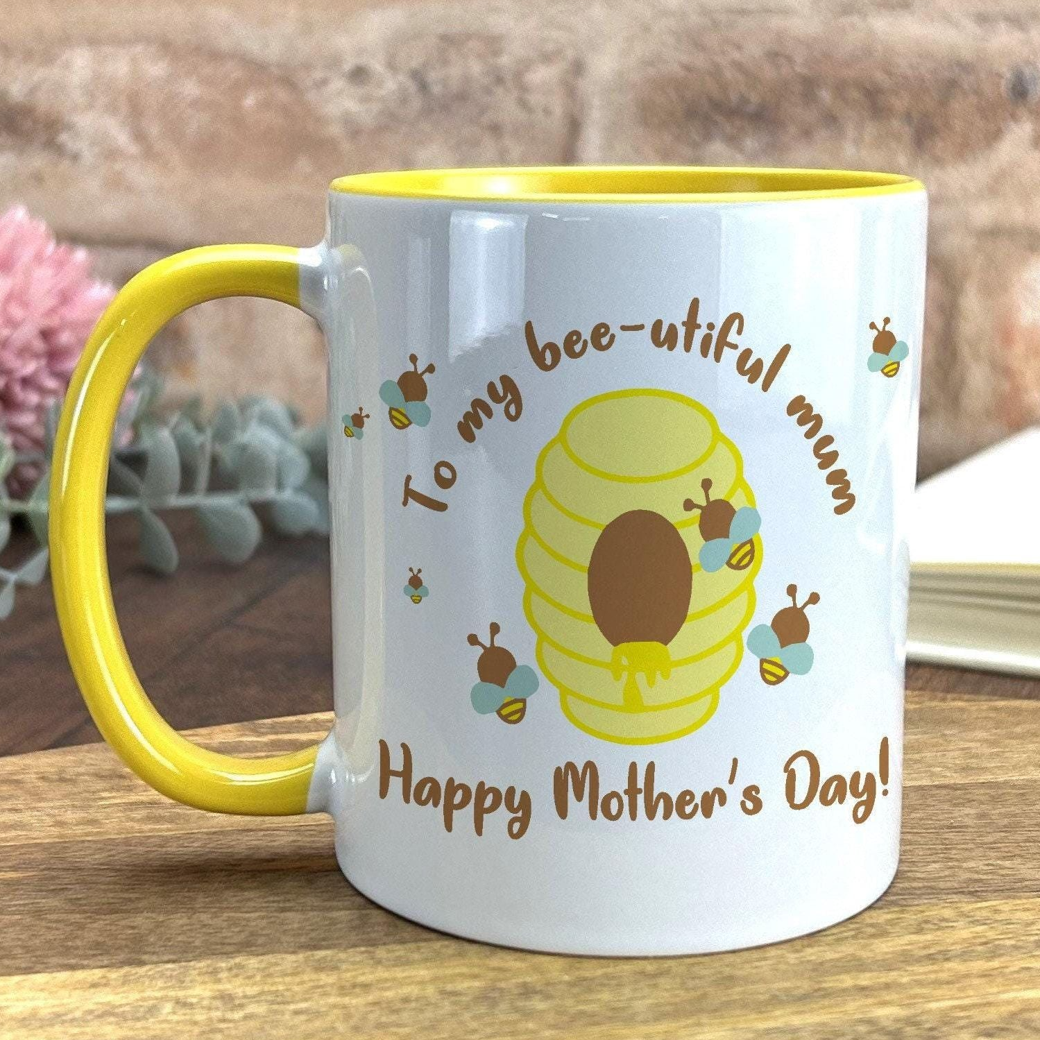 A white mug with a yellow rim, featuring a bee and honeycomb design, along with the text "To my bee-utiful mum Happy Mother's Day!"