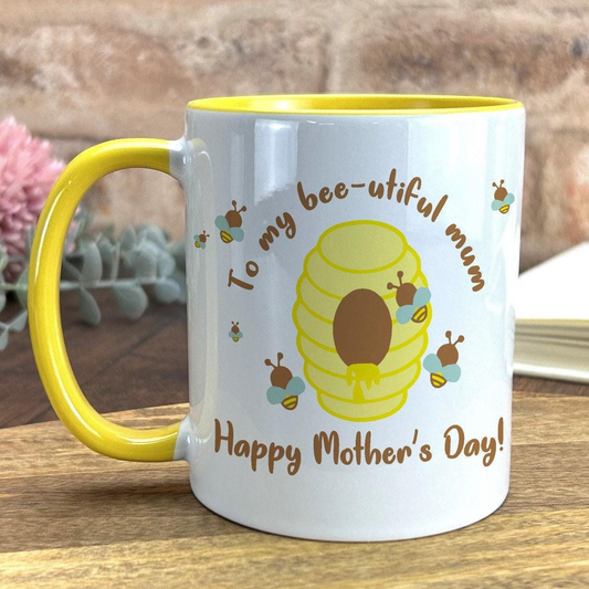 A white mug with a yellow rim, featuring a bee and honeycomb design, along with the text "To my bee-utiful mum Happy Mother's Day!"